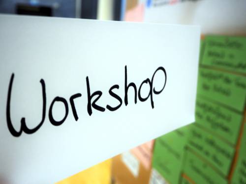 Auf einem weißen Kärtchen steht mit der Hand geschrieben "Workshop", dahinter ist eine Metaplanwand, an die weitere Kärtchen in anderen Farben geheftet sind.
