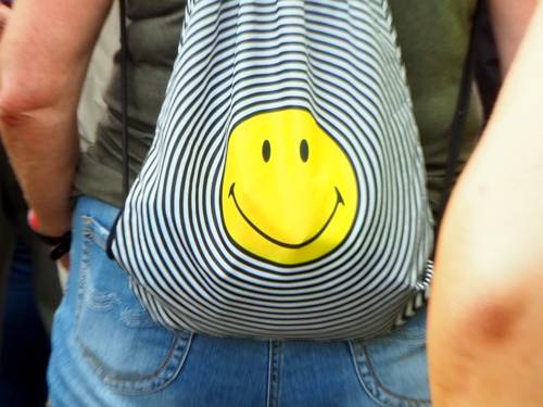 Eine Person trägt einen Turnbeutel auf dem Rücken und ist mit mehreren anderen Menschen unterwegs. Auf dem Turnbeutel ist ein lächelnder Smiley, umrahmt von größer werdenden Kreisen.