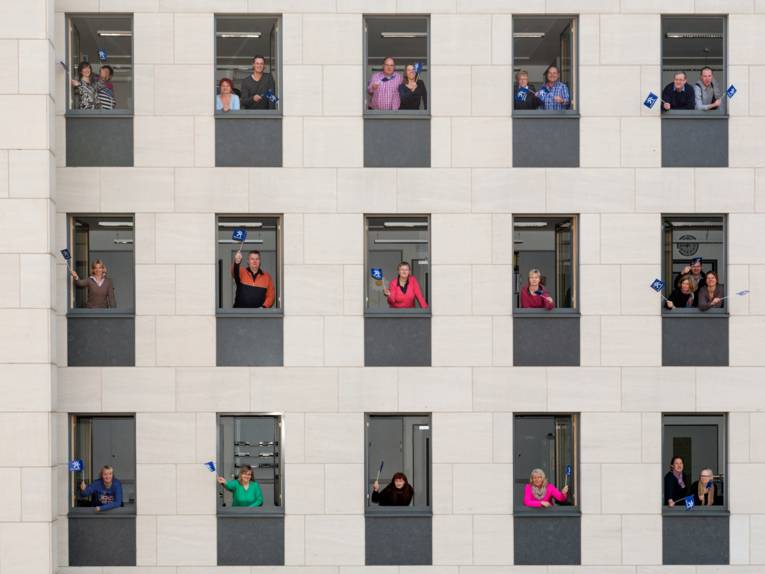 Beschäftigte schauen aus Bürofenstern und schwenken ein Regions-Fähnchen.