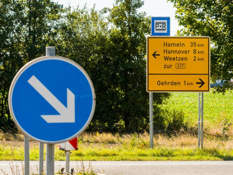 Im Vordergrund das runde Verkehrszeichen für vorgeschriebene Vorbeifahrt (rechts vorbei) mit einem weißen Pfeil auf blauem Grund, der nach rechts unten zeigt und im Hintergrund ein Wegweiser, der die Entfernung zu den Orten Hameln, Hannover, Weetzen und Gehrden zeigt.