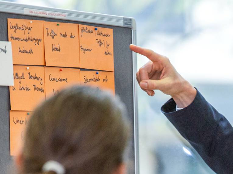 Eine Person schaut auf eine Metaplanwand. Dorthin zeigt eine andere Person mit dem Zeigefinger.