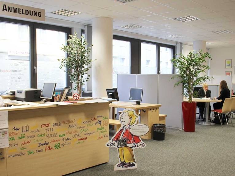 Im Vordergrund der Anmeldetresen im FamilienServiceBüro, im Hintergrund berät eine Mitarbeiterin eine junge Frau an einem Tisch.
