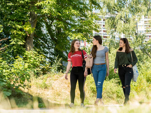 Dre Mädchen gehen durch eine Grünfläche, im Hintergrund sind Wohnhäuser für mehrere Parteien. Dre Mädchen gehen durch eine Grünfläche, im Hintergrund sind Wohnhäuser für mehrere Parteien.